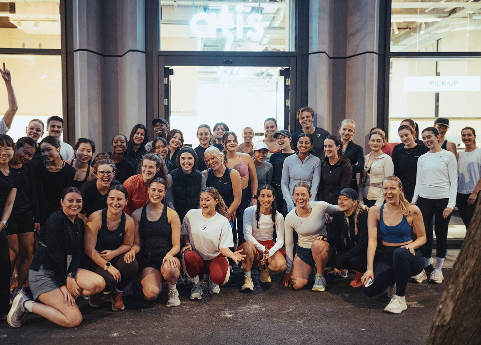 Large post‑run group smiling outside glowing atis storefront in London, celebrating community run