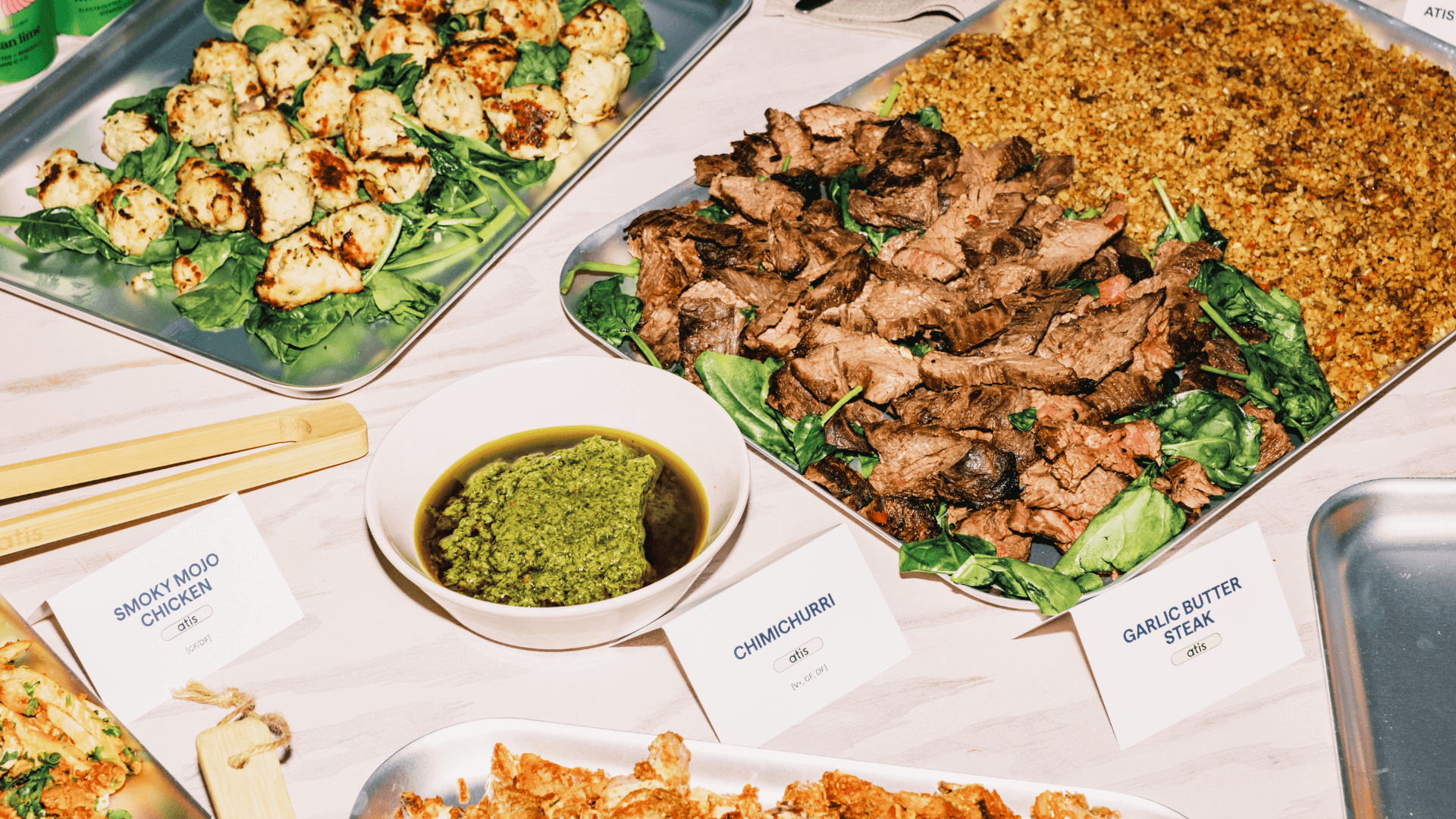 Overhead view of atis catering platters: garlic butter steak, smoky mojo cauliflower bites, chimichurri sauce and seasoned grains on a wooden table
