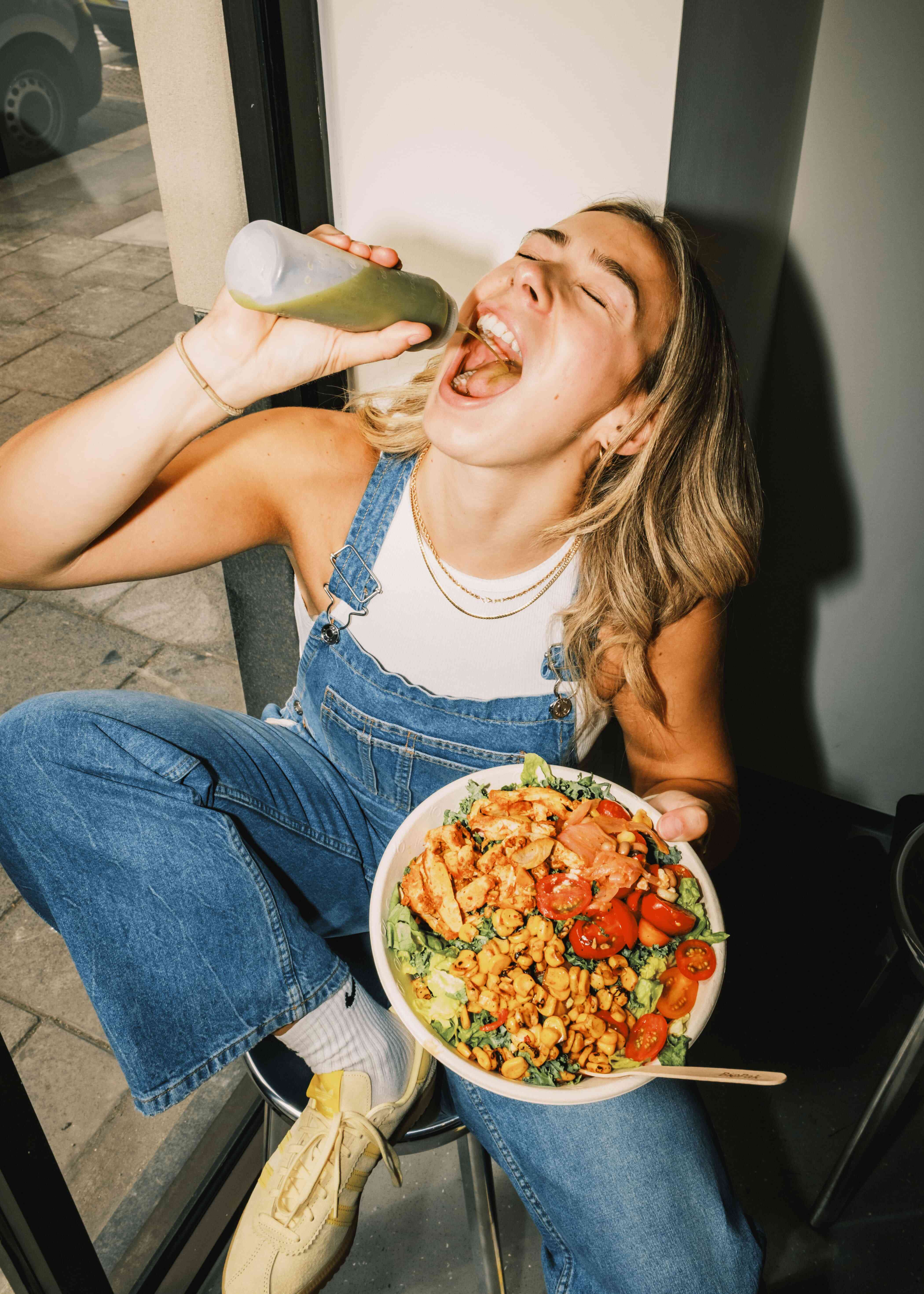 Playful customer sipping green dressing from bottle while holding atis chicken salad bowl by a window