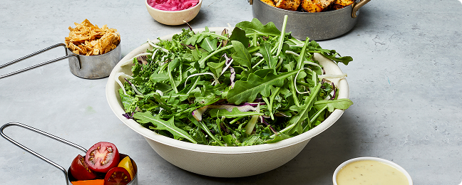 Overhead shot of atis build‑your‑own salad bowl with mixed kale and rocket leaves, surrounded by cups of toppings and dressing