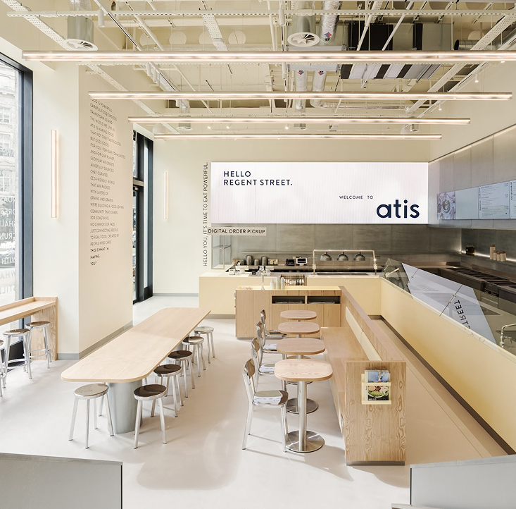 Bright, minimalist interior of atis Regent Street with pale wood communal tables, counter‑service prep line and large ‘Hello Regent Street. Welcome to atis’ backlit sign