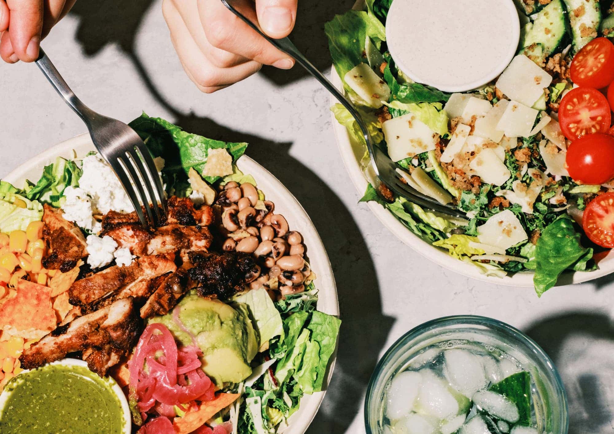 Two atis bowls on a table, one with blackened chicken, avocado, black eyed peas, pickled red onion, goats cheese and green dressing, the other a caesar-style salad with cherry tomatoes, parmesan and bread crumbs at an atis London restaurant
