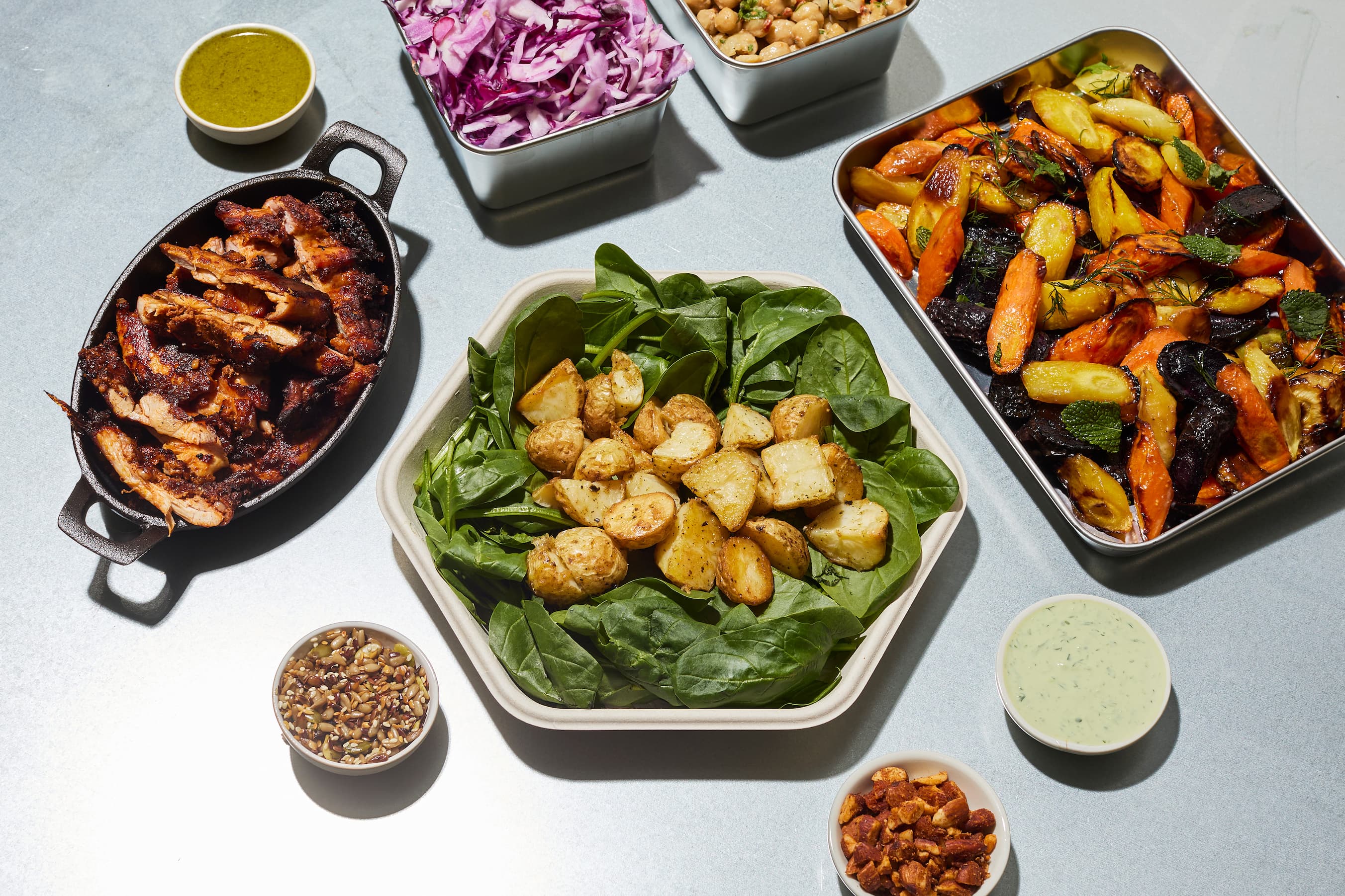 atis build your own plate spread showing blackened chicken, roasted potatoes, spinach, roasted carrots and beetroot, red cabbage, chickpeas, omega seeds, herb dressing and green sauce on a menu table