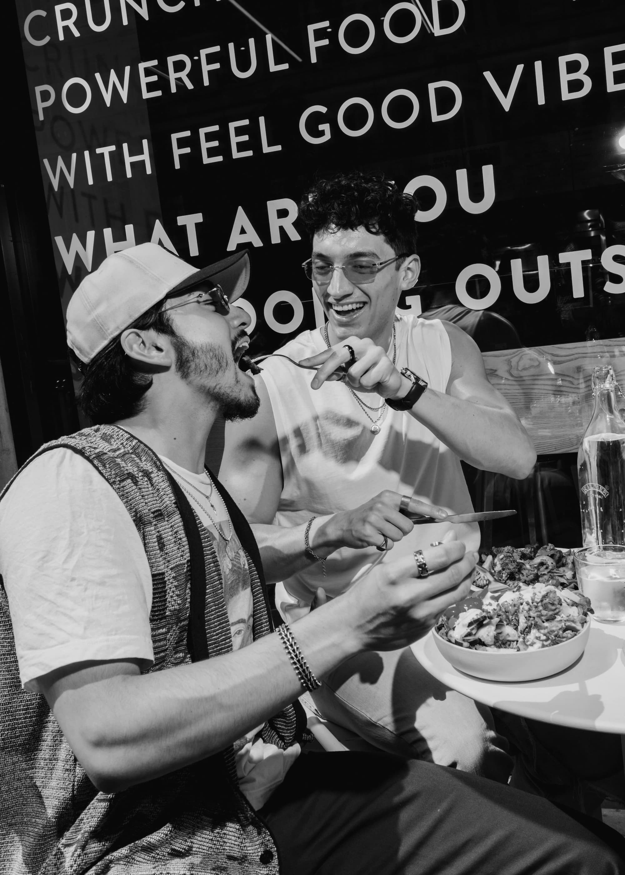 Two friends laughing and sharing an atis bowl outside an atis London restaurant with the storefront window copy visible in the background