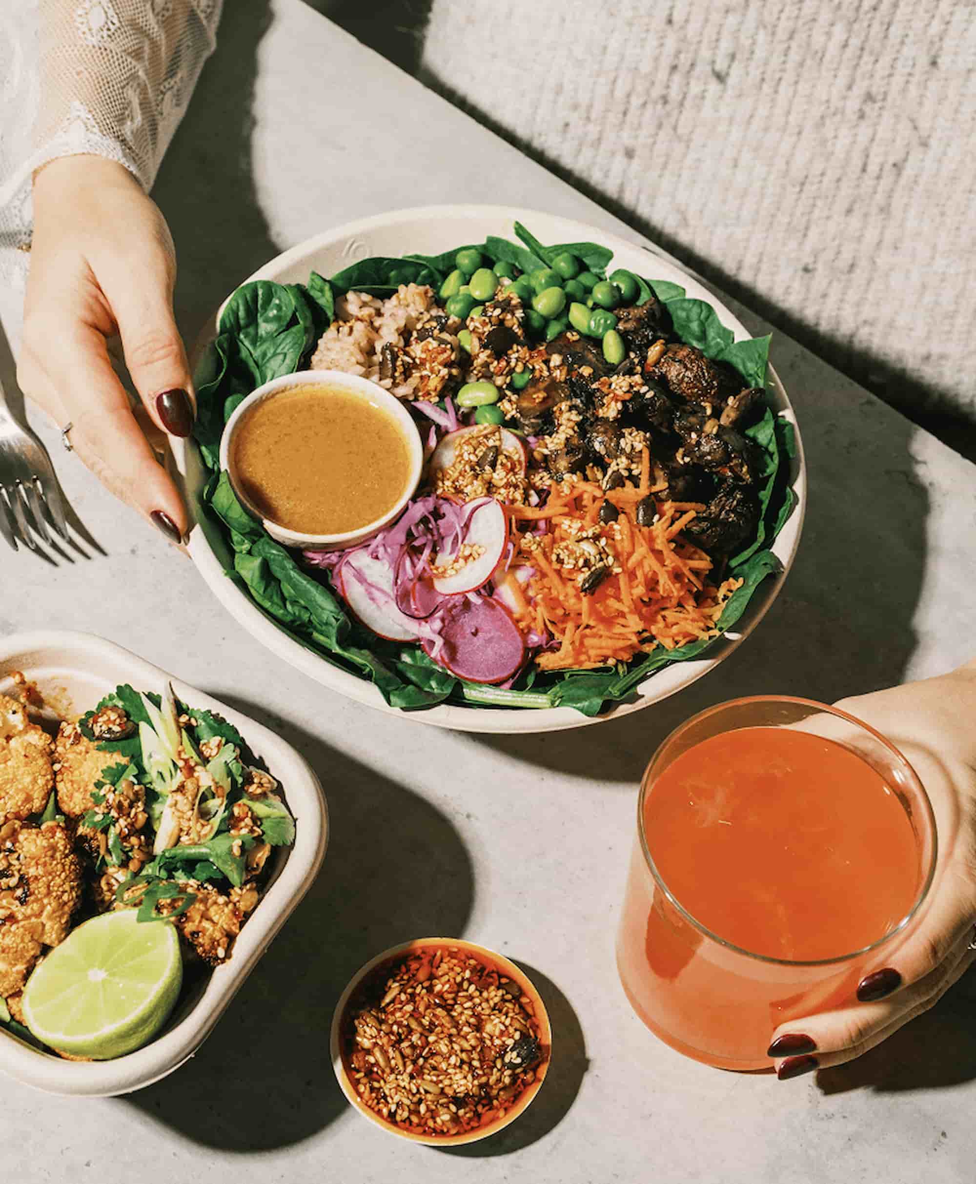 atis bowl with roasted mushrooms, edamame, shredded carrot, pickled red onion, brown rice, spinach and miso dressing alongside a crispy cauliflower side and fresh juice at an atis London restaurant