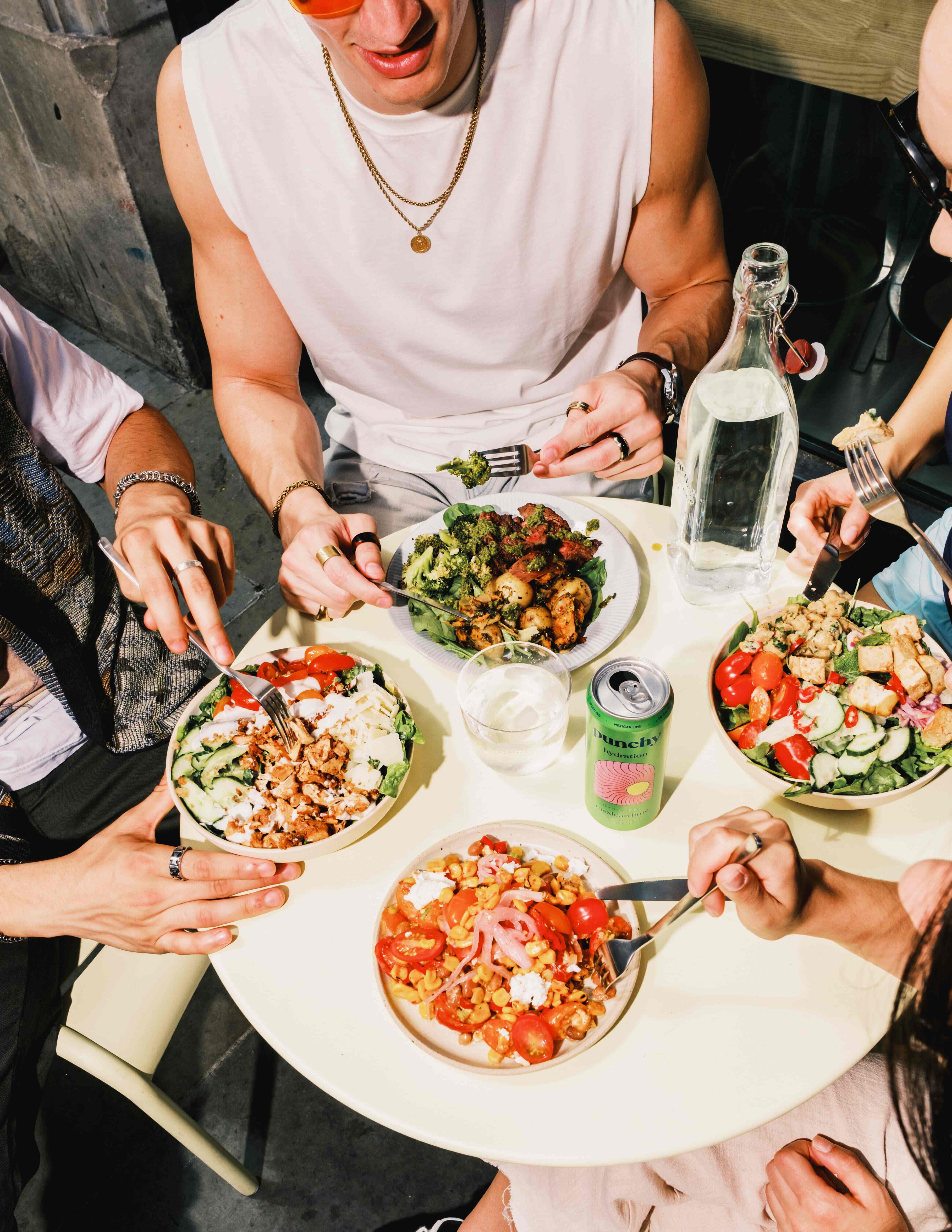 Group of friends enjoying colourful atis salad bowls and canned drinks around a sunny outdoor table in London