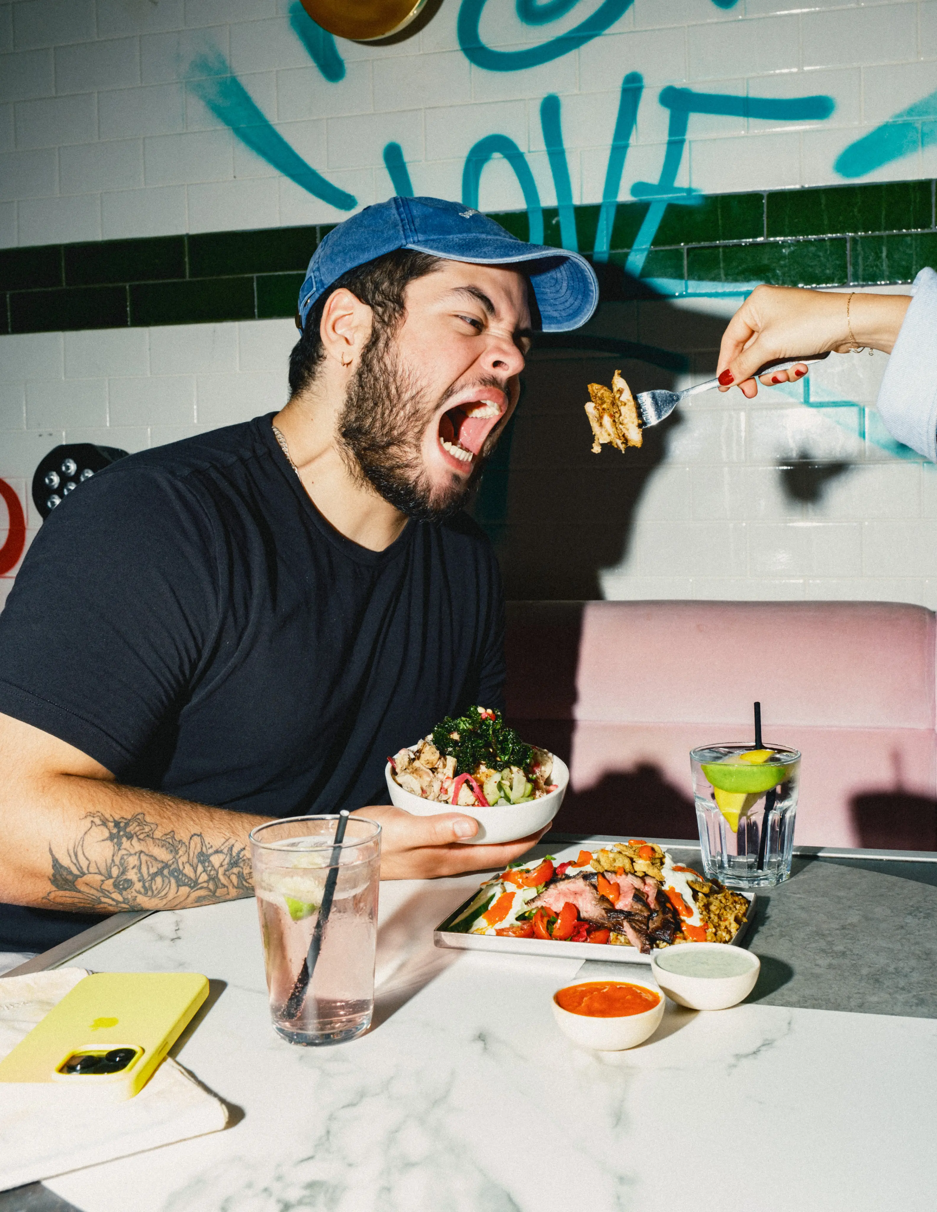 Bearded man wearing an atis cap, opening wide as a friend feeds him a forkful of atis salad bowl at a marble‑topped table”