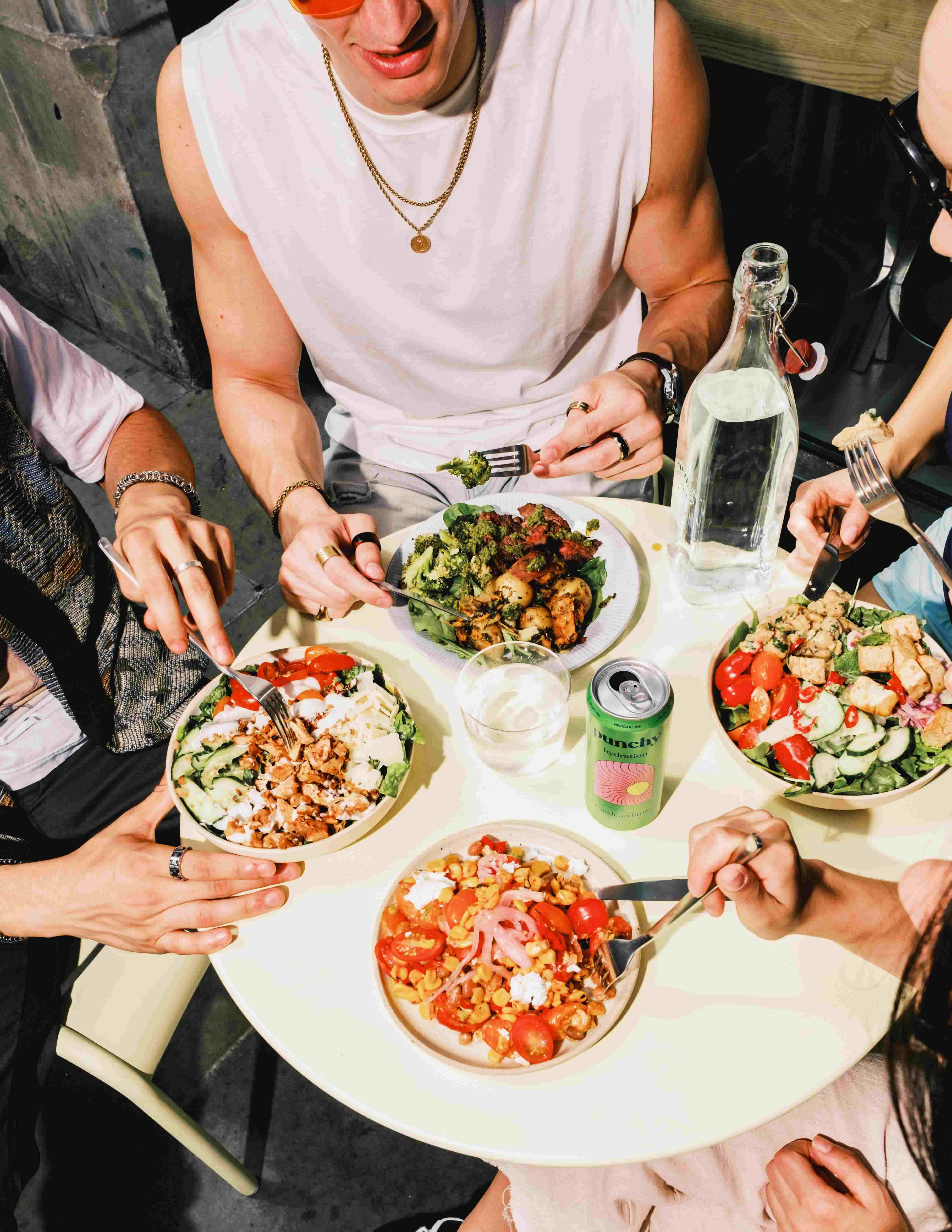 Group of friends enjoying colourful atis salad bowls and canned drinks around a sunny outdoor table in London