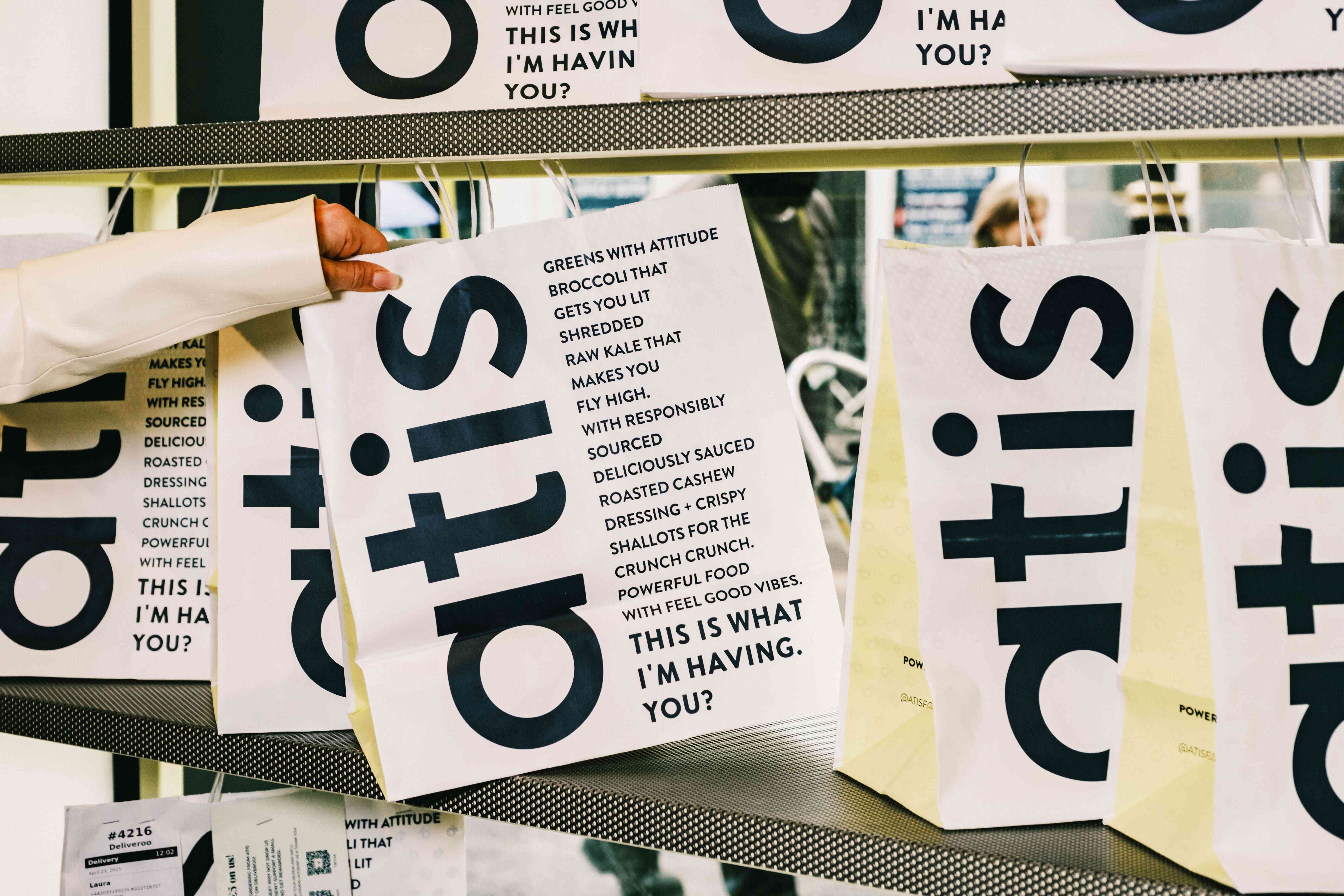Close‑up of atis branded paper bags hanging on a metal rack, featuring bold navy logo and menu‑style text about greens, cashew dressing and ‘This is what I’m having. You?’”