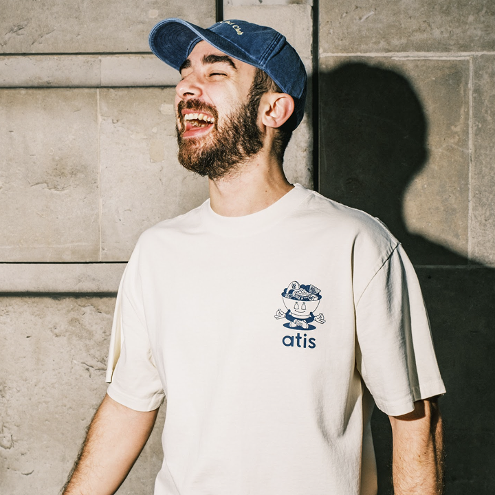 Bearded man laughing while wearing an off‑white atis T‑shirt printed with a cartoon salad‑bowl mascot