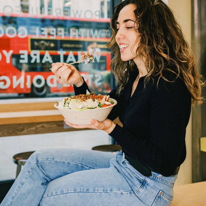 Woman eating an atis salad bowl with romaine, cherry tomatoes and granola topping inside an atis London restaurant with a red London bus visible through the window